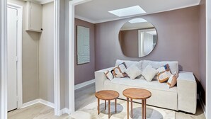 Een moderne woonkamer met een bank, twee ronde houten tafels, een grote spiegel en een wanddecoratie met geometrische vormen.