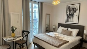 A hotel room with a bed, a desk with a chair, a small table with a vase, a mirror, and a framed picture on the wall. - Charming Apartment Center of Paris (Paris)
