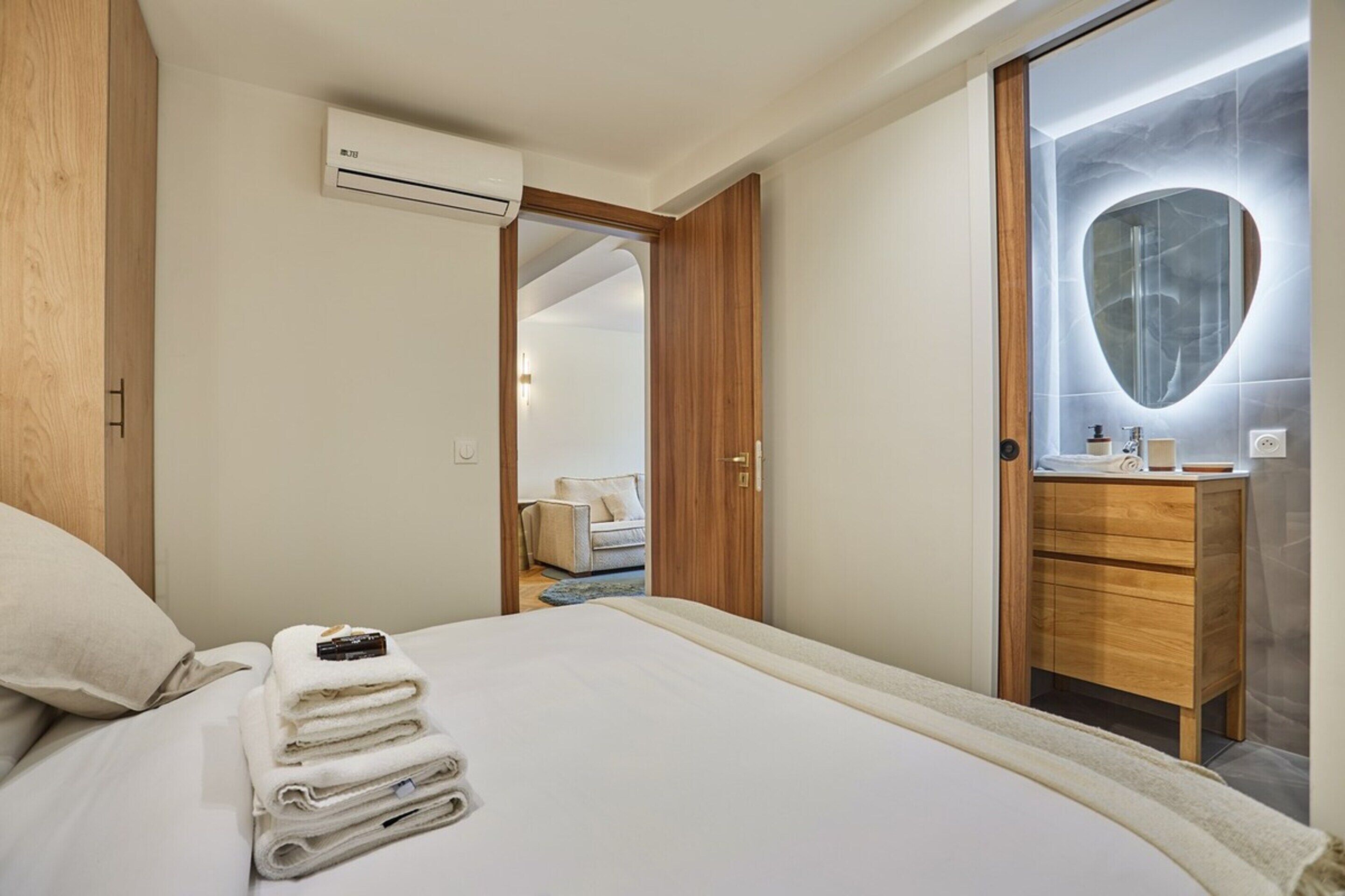 A hotel room with a bed, a wooden wardrobe, a bathroom with a marble vanity, and an air conditioning unit.