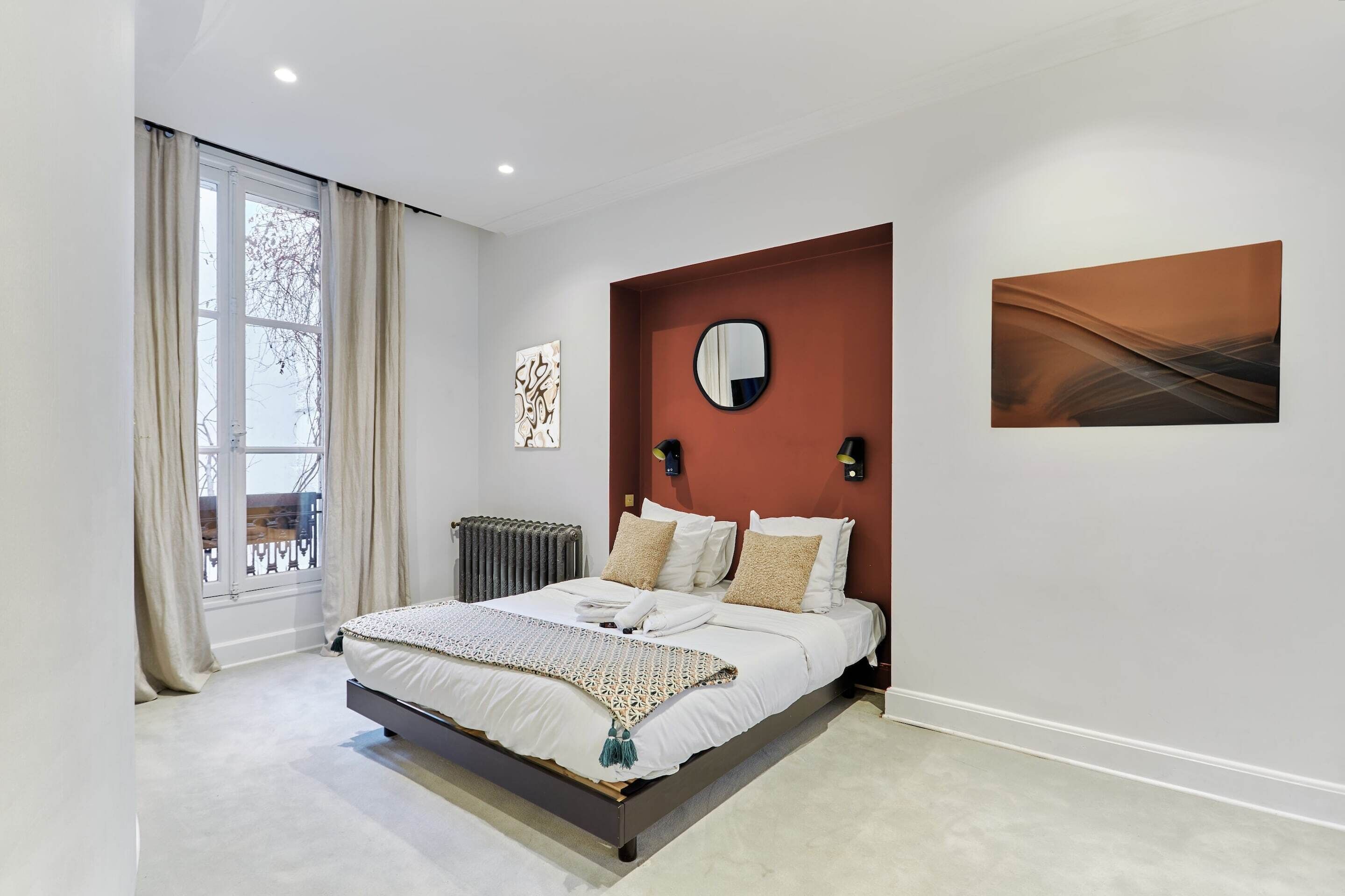 A modern bedroom with a large bed, a radiator, a window with curtains, and a wall-mounted mirror.