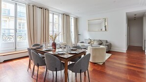 A modern dining area with a wooden table set for four, a sofa, and a large window with curtains. - Sublime 3 Bedroom Flat Palais Royal Bourse (Paris)