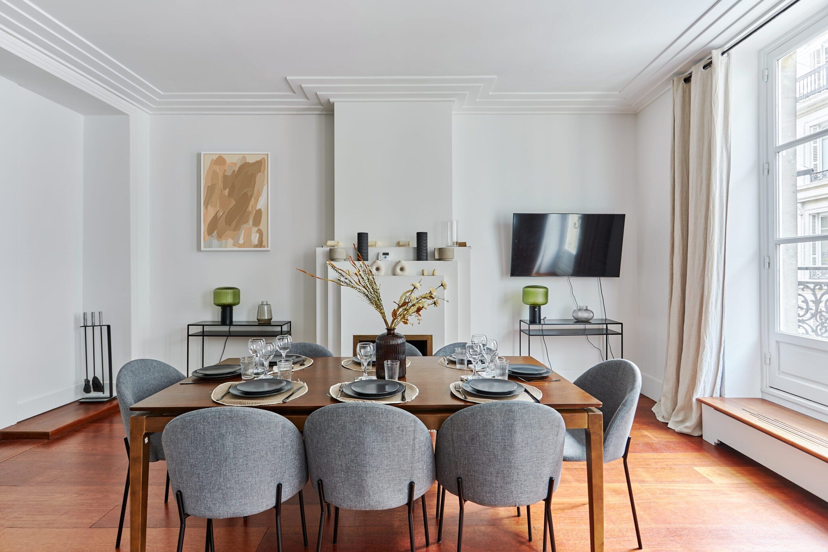 A modern dining room with a wooden table set for four, grey upholstered chairs, a vase with dried flowers, and a flat-screen TV mounted on the wall.