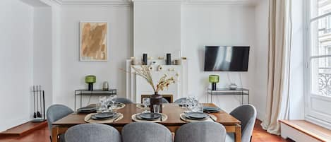 A modern dining room with a wooden table set for four, grey upholstered chairs, a vase with dried flowers, and a flat-screen TV mounted on the wall.