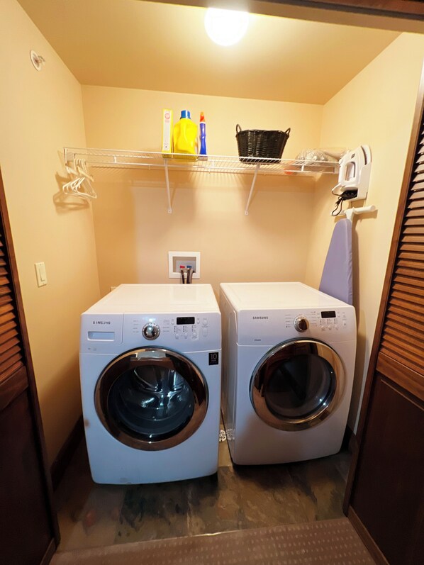 A laundry room with two front-loading washing machines, a shelf with cleaning supplies, and a hanging rack.