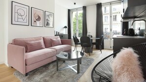 Una sala de estar moderna con un sofá rosa, una mesa de centro de vidrio y una silla negra. Hay obras de arte enmarcadas en la pared, un área de comedor con sillas y vistas de la ciudad a través de amplios ventanales.