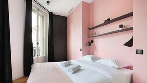 Una cama bien tendida con sábanas blancas y toallas enrolladas, una pared rosa y una ventana con cortinas negras.