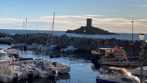 Un puerto deportivo con barcos amarrados, un rompeolas rocoso y una torre en una pequeña isla al fondo.