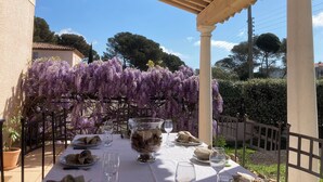 Un área de comedor al aire libre con una mesa dispuesta para dos personas, rodeada por una pérgola y glicinas moradas.