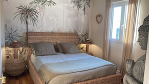Un dormitorio con cabecera trenzada, cama con edredón blanco, dos lámparas de noche, una ventana con cortinas y un mural de plantas tropicales.