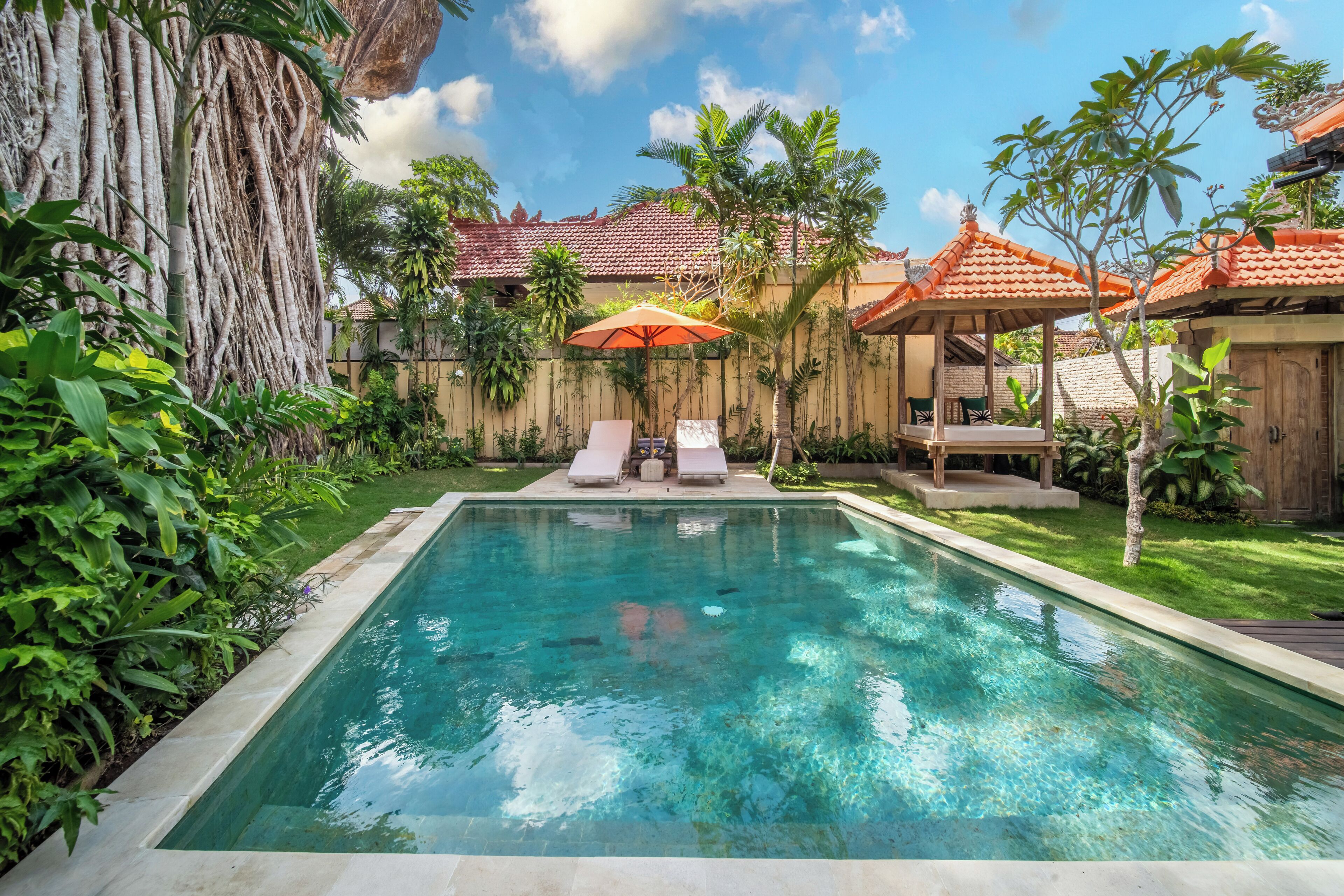 A rectangular pool with clear water, surrounded by lush greenery and a wooden fence. There are lounge chairs and an umbrella under a gazebo.