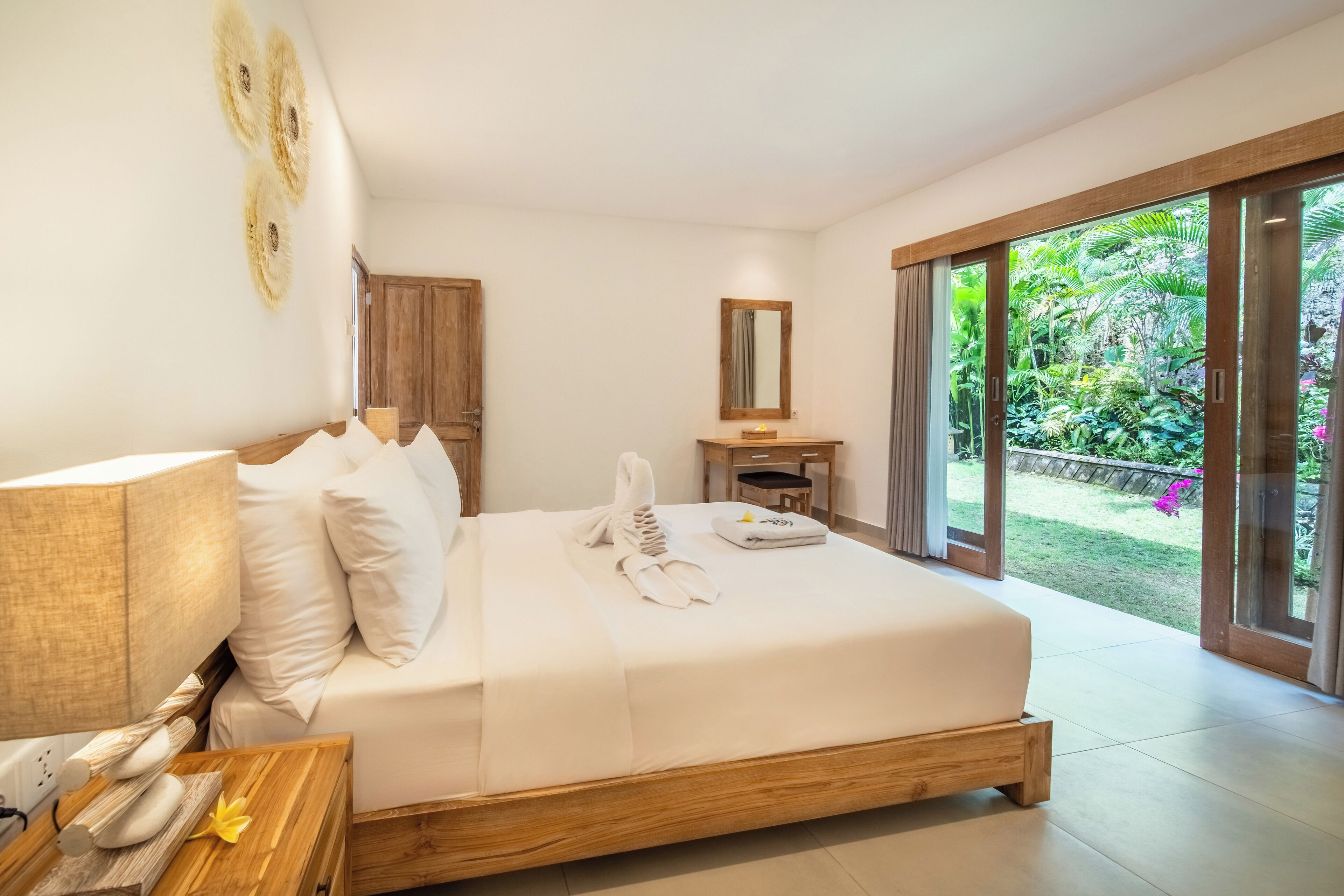 Een moderne hotelkamer met een groot bed, een houten nachtkastje, een spiegel en uitzicht op een weelderige tuin door de schuifglazen deur.