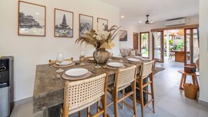 A dining area with a large table set for four, surrounded by wicker chairs. There are framed pictures on the wall, a vase with dried flowers, and a ceiling fan. - Tropical Villa Sinar Maluku (Ungasan)