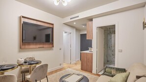 Una sala de estar moderna con un televisor de pantalla plana, una zona de comedor con mesas redondas, un sofá y una cocina al fondo.