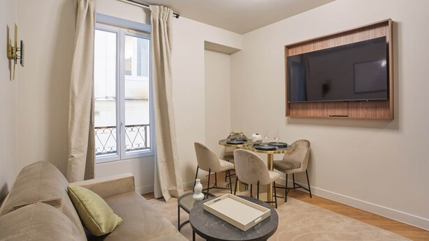 Una sala de estar con sofá, una mesa de comedor con sillas, televisor montado en la pared y ventana con cortinas.