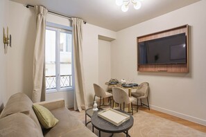 Ein Wohnzimmer mit einer Couch, einem Esstisch mit Stühlen, einem an der Wand befestigten Fernseher und einem Fenster mit Vorhängen.