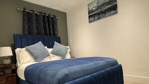 A bedroom with a large bed, dark curtains, a nightstand with a lamp, and a framed picture on the wall. - Deluxe 4 Bedroom House with Free Parking (Slough)