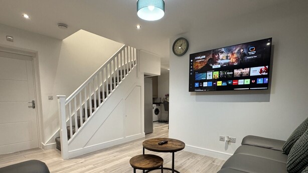 A modern living room with a staircase, a flat-screen TV mounted on the wall, and a grey sofa. - Deluxe 4 Bedroom House with Free Parking (Slough)