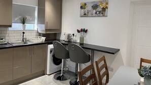 A compact kitchen with a built-in washing machine, two bar stools, and a dining table with chairs. - Deluxe 4 Bedroom House with Free Parking (Slough)