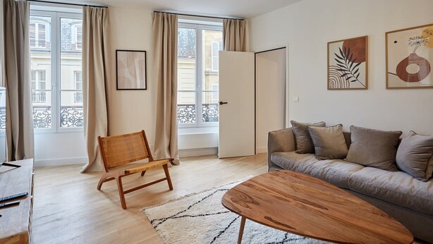 Ein modernes Wohnzimmer mit einer Couch, einem Holztisch, einem Korbstuhl und einem großen Fenster mit Vorhängen.
