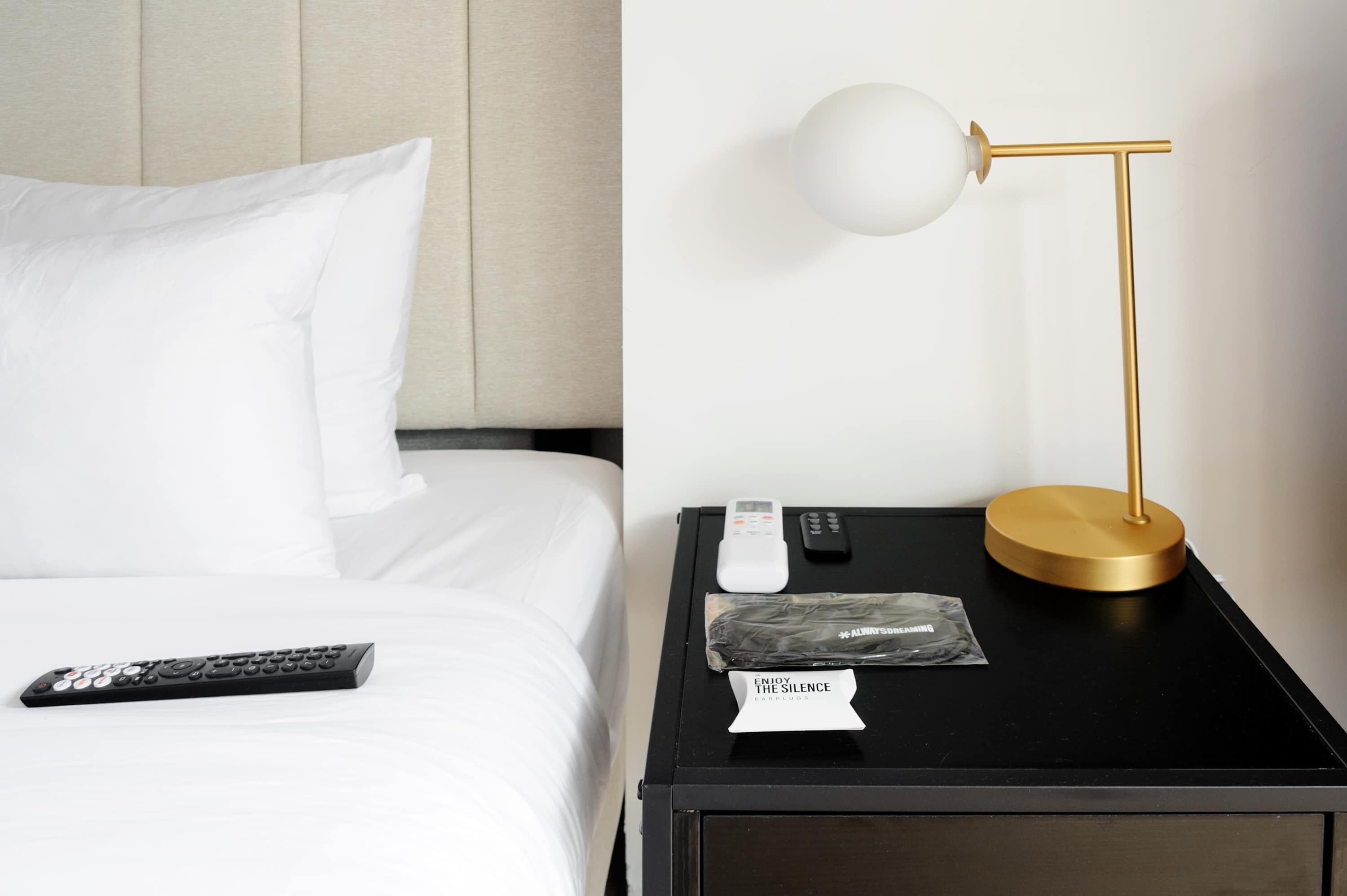 Una cama bien hecha con ropa blanca, un control remoto negro, una lámpara de noche blanca, un control remoto y un letrero que dice 'ENJOY THE SILENCE'.