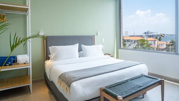 A hotel room with a large bed, a view of the city, and a wooden bench.