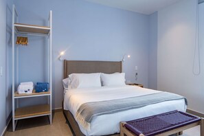 Een net opgemaakt bed met witte lakens, een houten bankje aan het voeteneind, een witte kledingkast met planken, en een wandlamp.