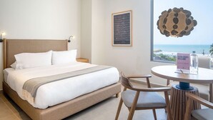 A hotel room with a bed, a round table with chairs, a framed artwork on the wall, and a view of the ocean.