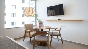 A modern living room with a round wooden table, two chairs, a TV mounted on the wall, and a large window with a view of buildings.