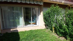 A residential building with a sliding glass door entrance, a small garden area with grass and a bush, and a wooden fence.