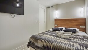 Ein Schlafzimmer mit einem großen Flachbildfernseher an der Wand, einem Holzkopfende und einem ordentlich bezogenen Bett mit einem gemusterten Überwurf.