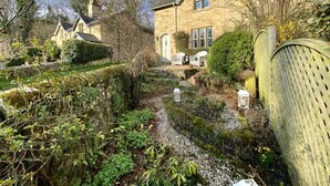 A stone house with a garden path leading to the entrance, surrounded by greenery and a wooden fence.