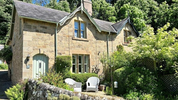 A stone house with a gabled roof, a front door, and two windows. There are two wicker chairs outside.