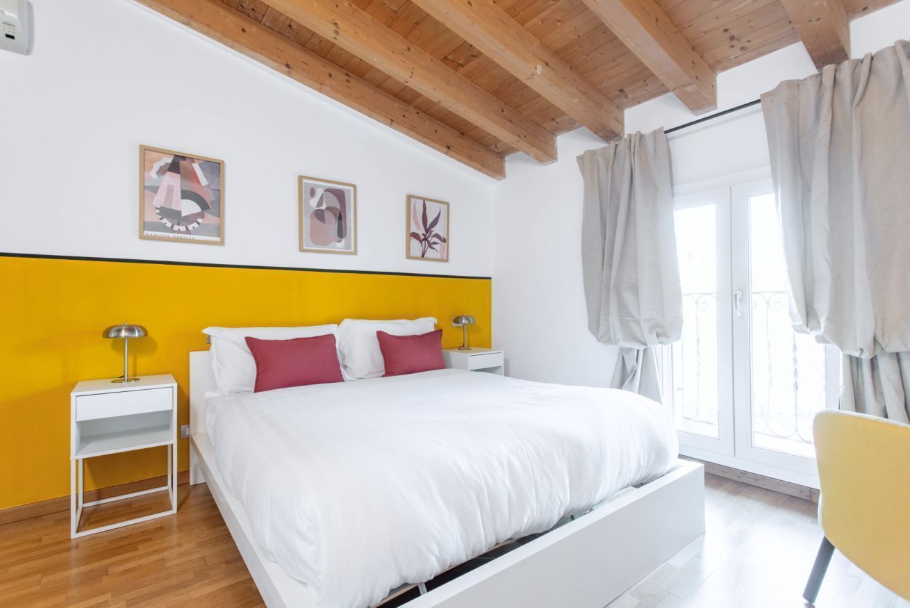 A modern bedroom with a white bed, yellow accent wall, and wooden ceiling beams.