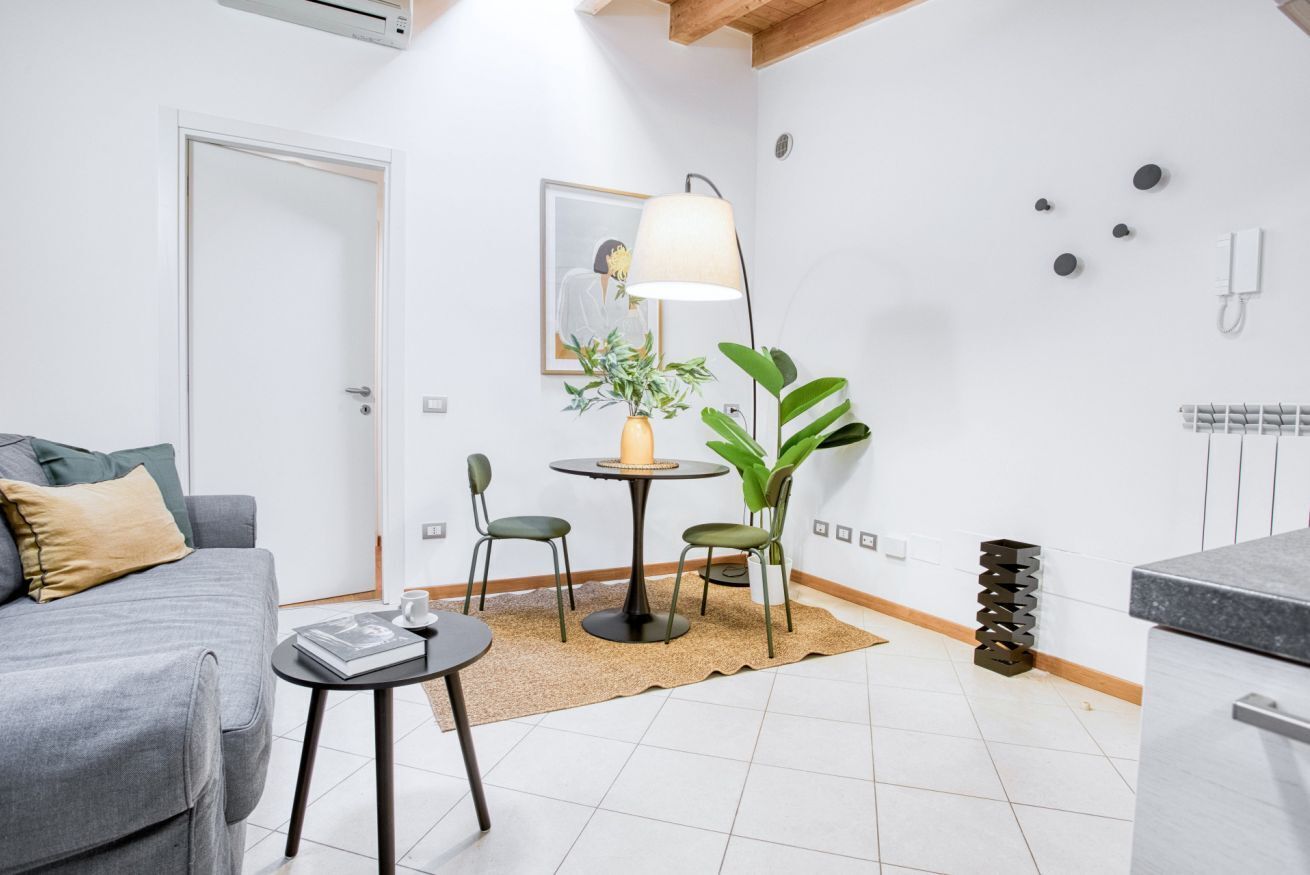 A modern living room with a grey sofa, a small round table with a book, a floor lamp, and a potted plant.