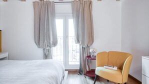 A bedroom with a bed, a chair, a small table with a book, and a window with curtains.
