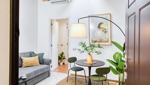 A compact living space with a grey sofa, a small round table with chairs, a lamp, and a potted plant.