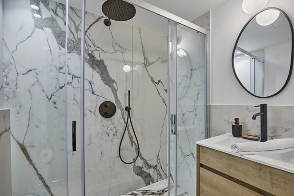 A modern bathroom with a glass-enclosed shower, marble walls, a wooden vanity, and a round mirror.
