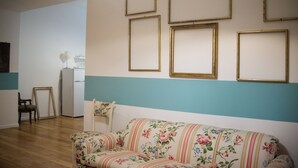A living room with a floral sofa, a white armchair, and a small table with a lamp. There are several empty picture frames on the wall. - Cheerful little house in the heart of the historic center  (Palermo)