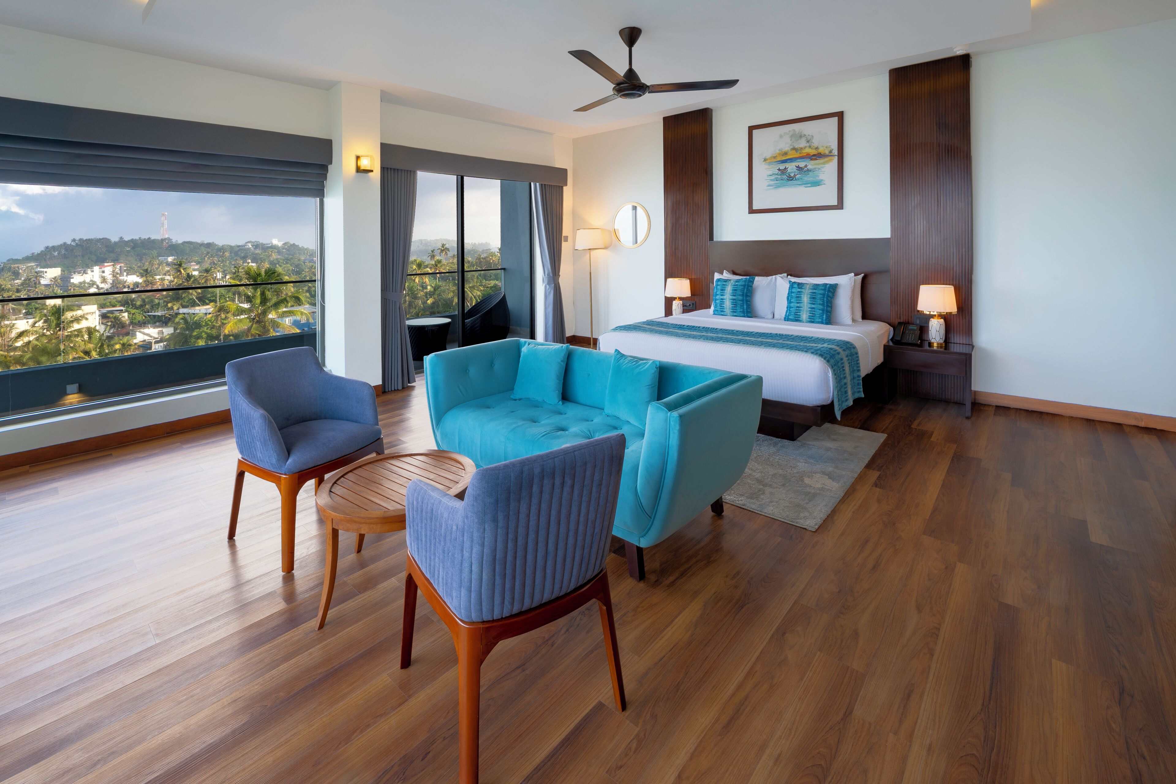 A modern hotel room with a large bed, a seating area with two chairs, a ceiling fan, and a view of a cityscape through large windows.