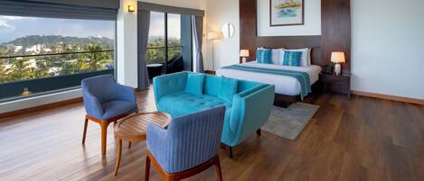 A modern hotel room with a large bed, a seating area with two chairs, a ceiling fan, and a view of a cityscape through large windows.