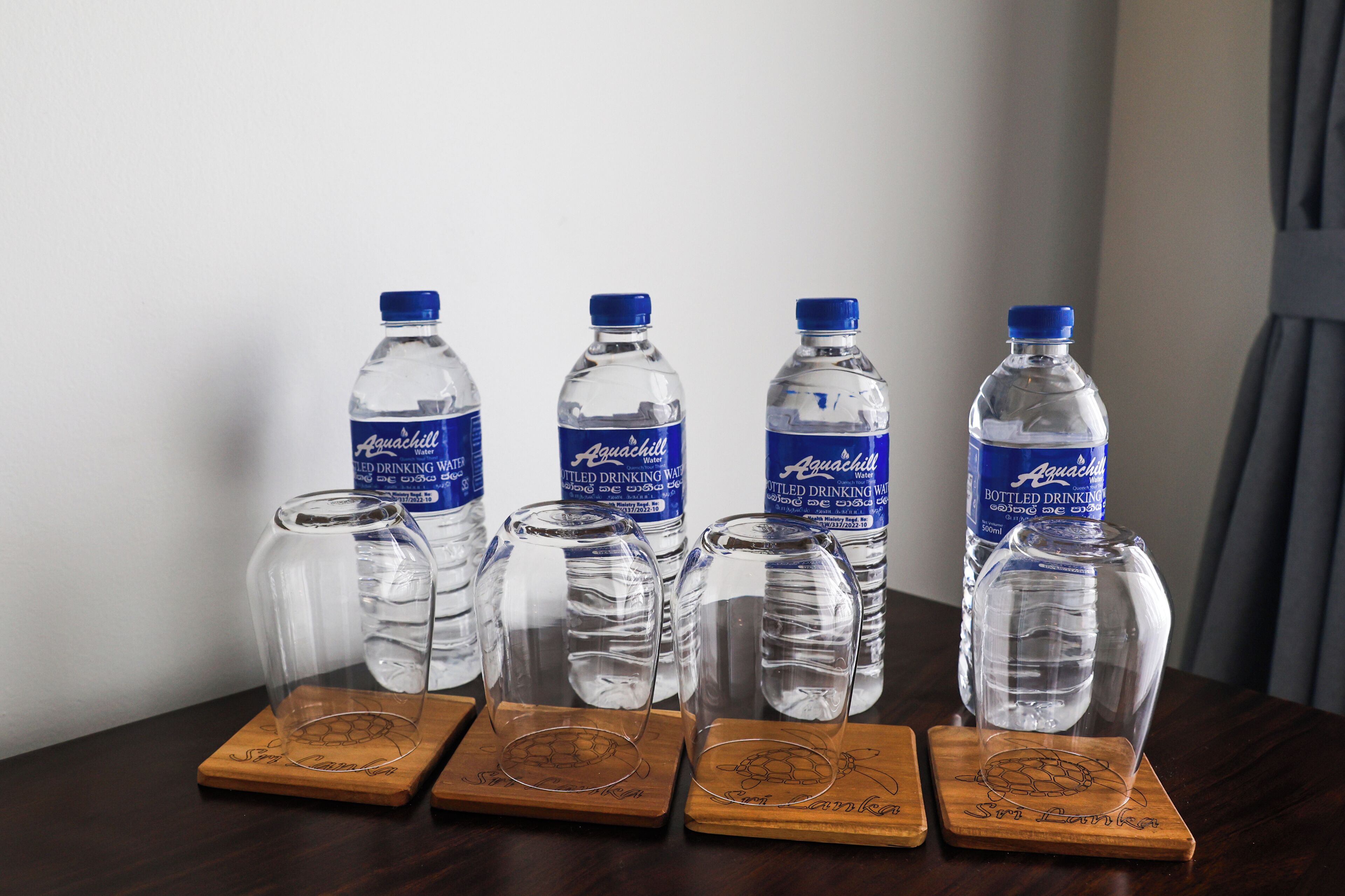 Una mesa con seis botellas de agua Aquashill y cuatro vasos en posavasos de madera.