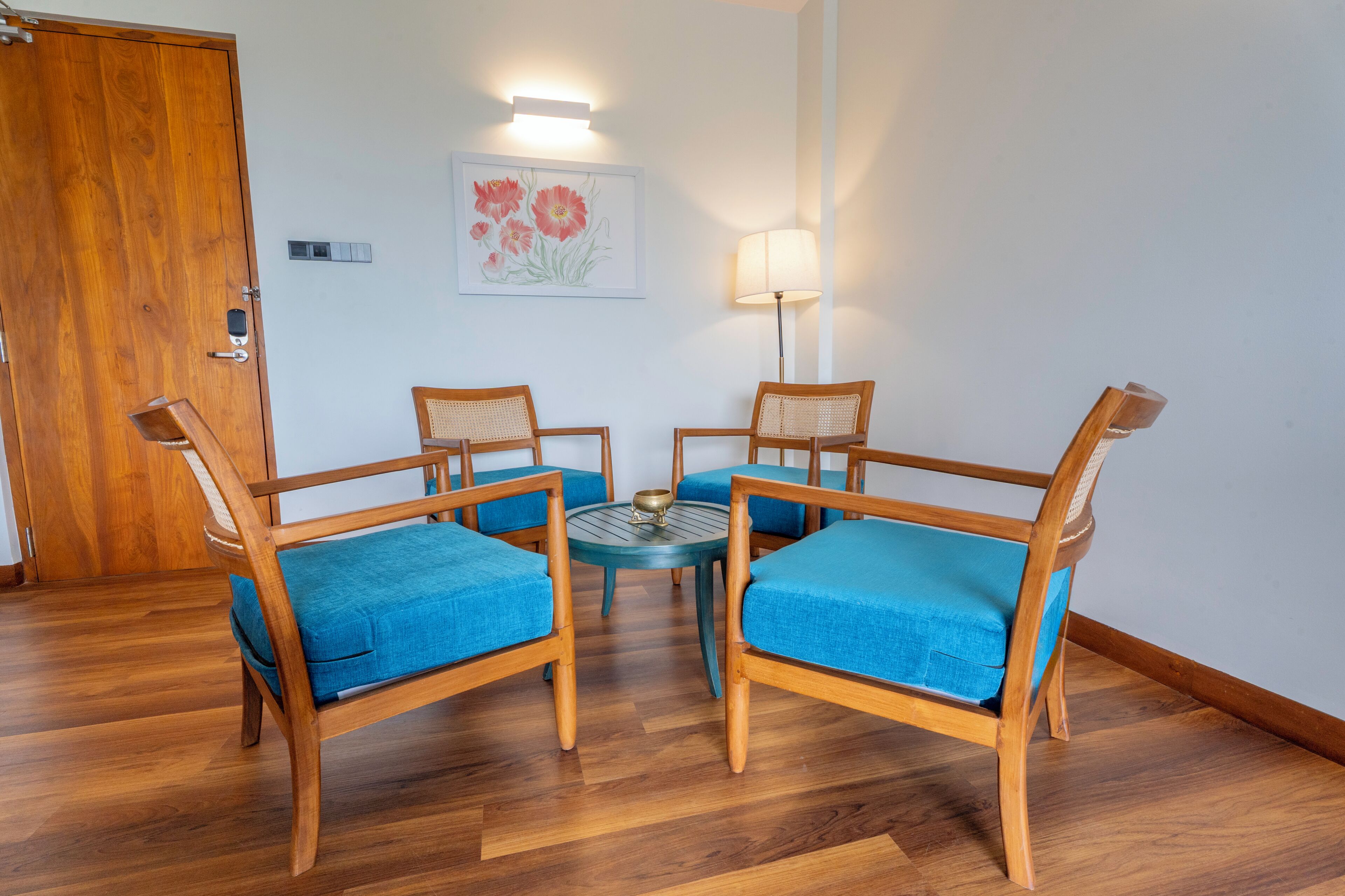 Une pièce avec une porte en bois, une œuvre d’art florale encadrée, une table ronde avec des chaises et un lampadaire.