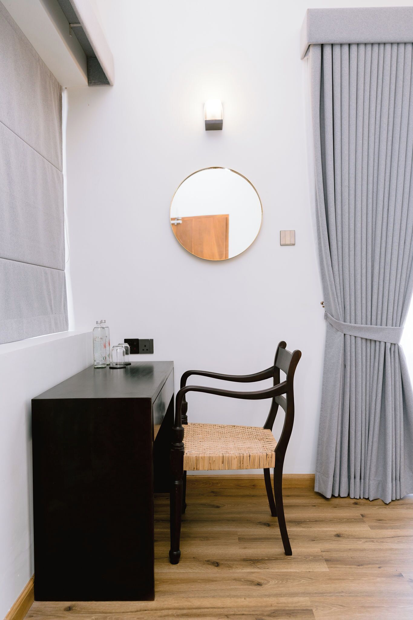 Une pièce avec un bureau en bois foncé, un miroir rond, une chaise en osier, un rideau gris et un interrupteur sur le mur.