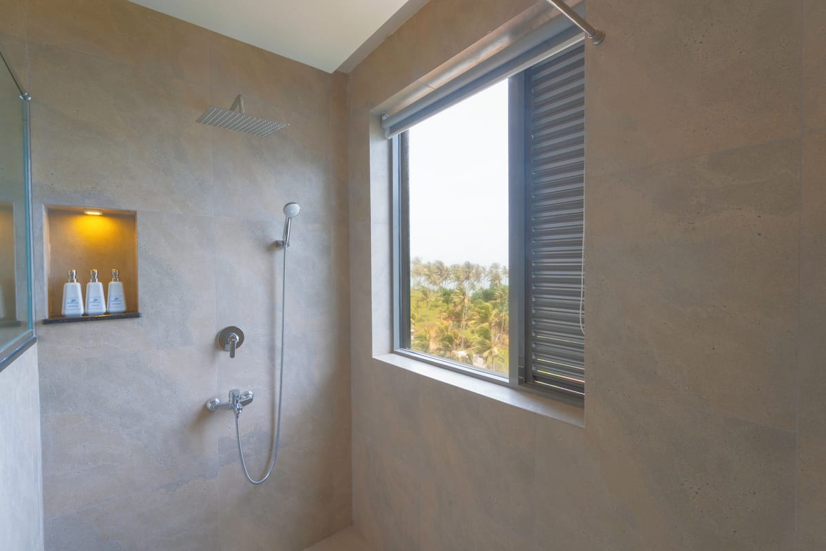 A bathroom with a shower, a window with blinds, and a shelf with toiletries.
