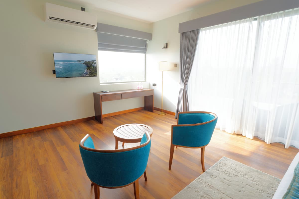 A room with a wooden floor, a desk with a computer, two blue chairs, a TV on the wall, and a window with curtains.