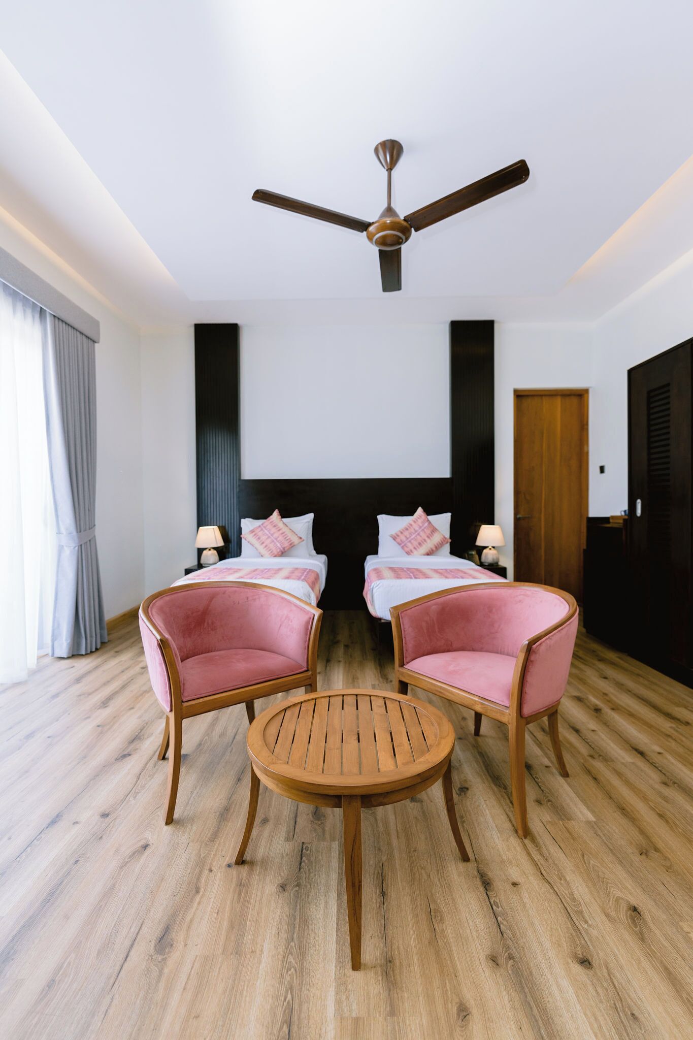 A hotel room with a bed, two pink chairs, a round wooden table, a ceiling fan, and a large window with curtains.