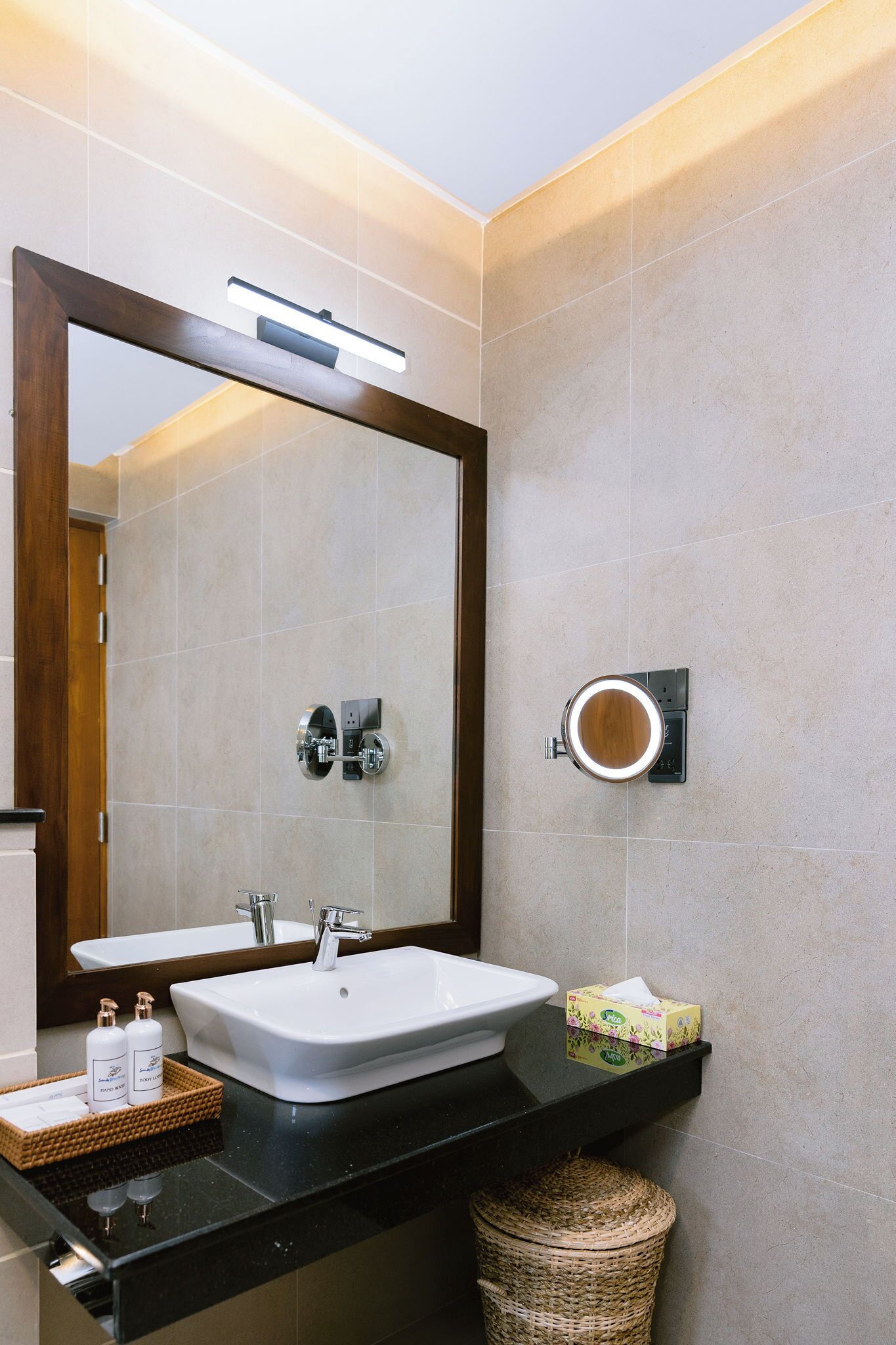 A bathroom with a large mirror, a white sink, a black countertop, a wicker basket, and a small tissue box.
