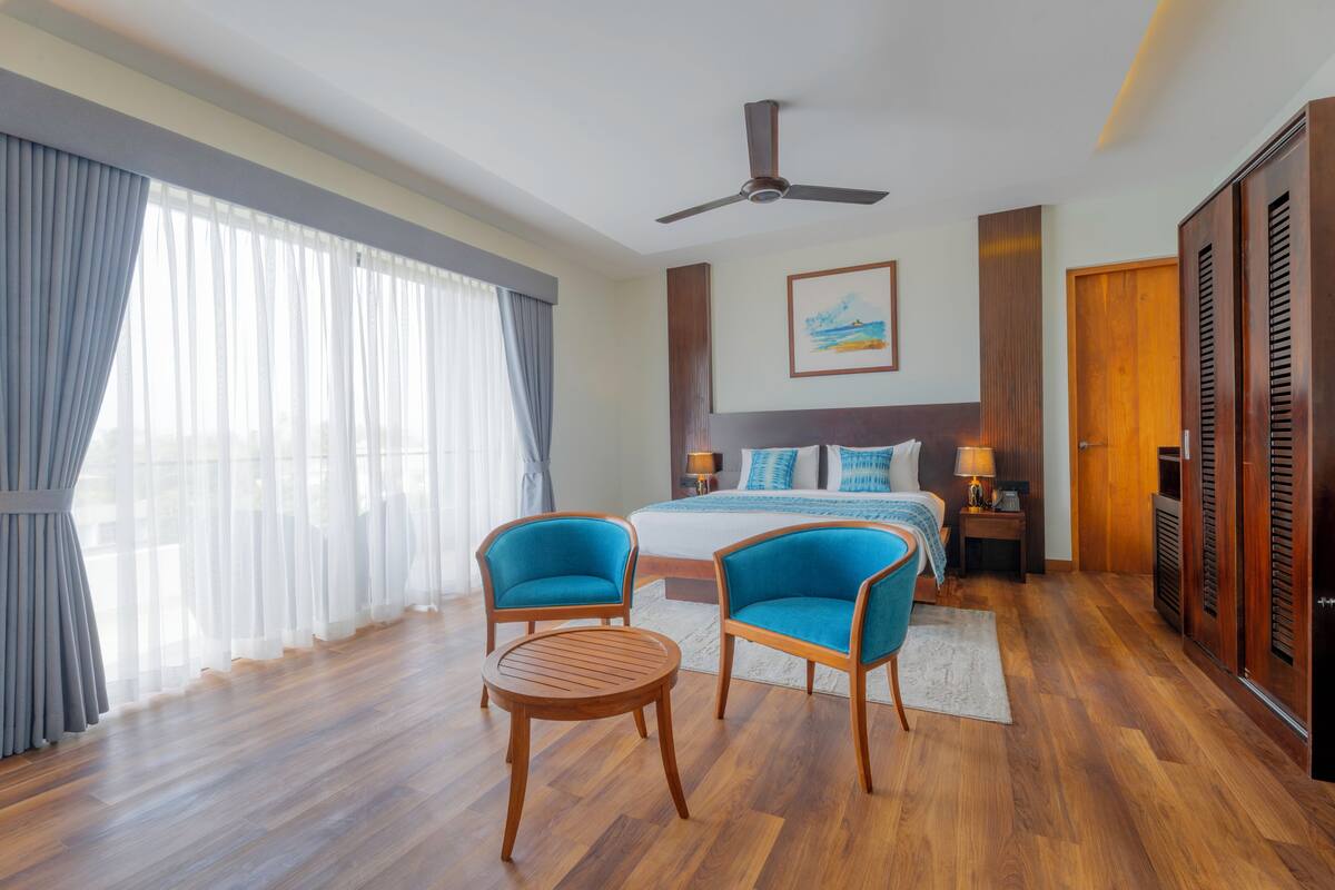 A hotel room with a bed, two blue armchairs, a round wooden table, a ceiling fan, and wooden wardrobes.