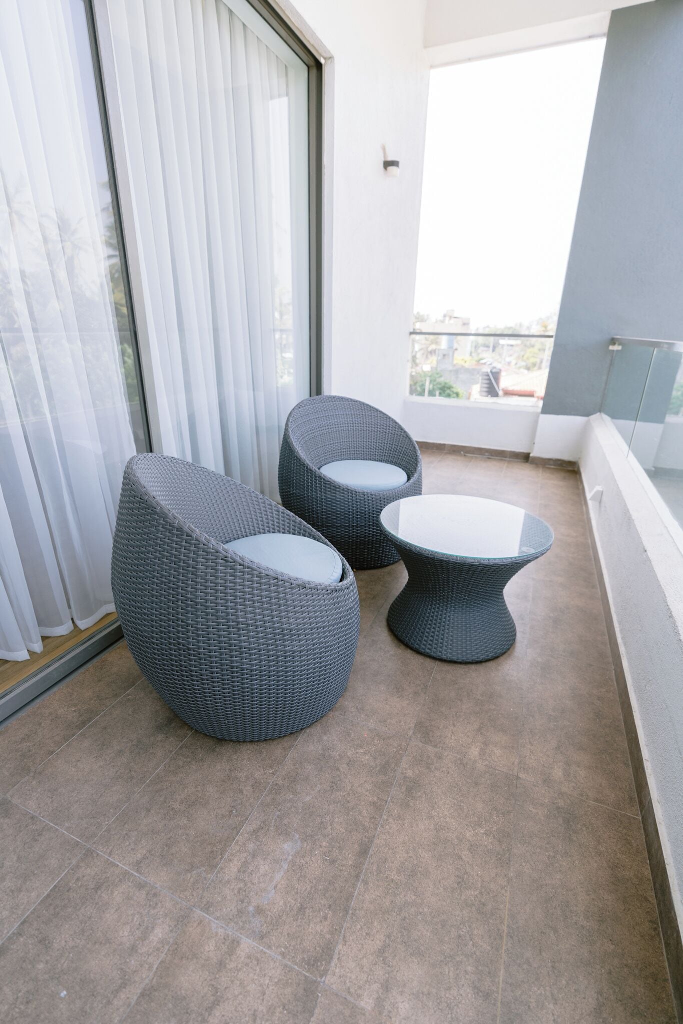 A balcony with two wicker chairs and a small table, a sliding glass door, and a view of buildings.
