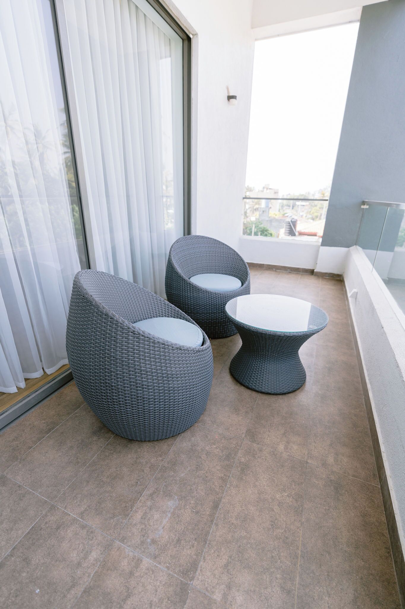 A balcony with two wicker chairs and a small table, a sliding glass door, and a view of the city.